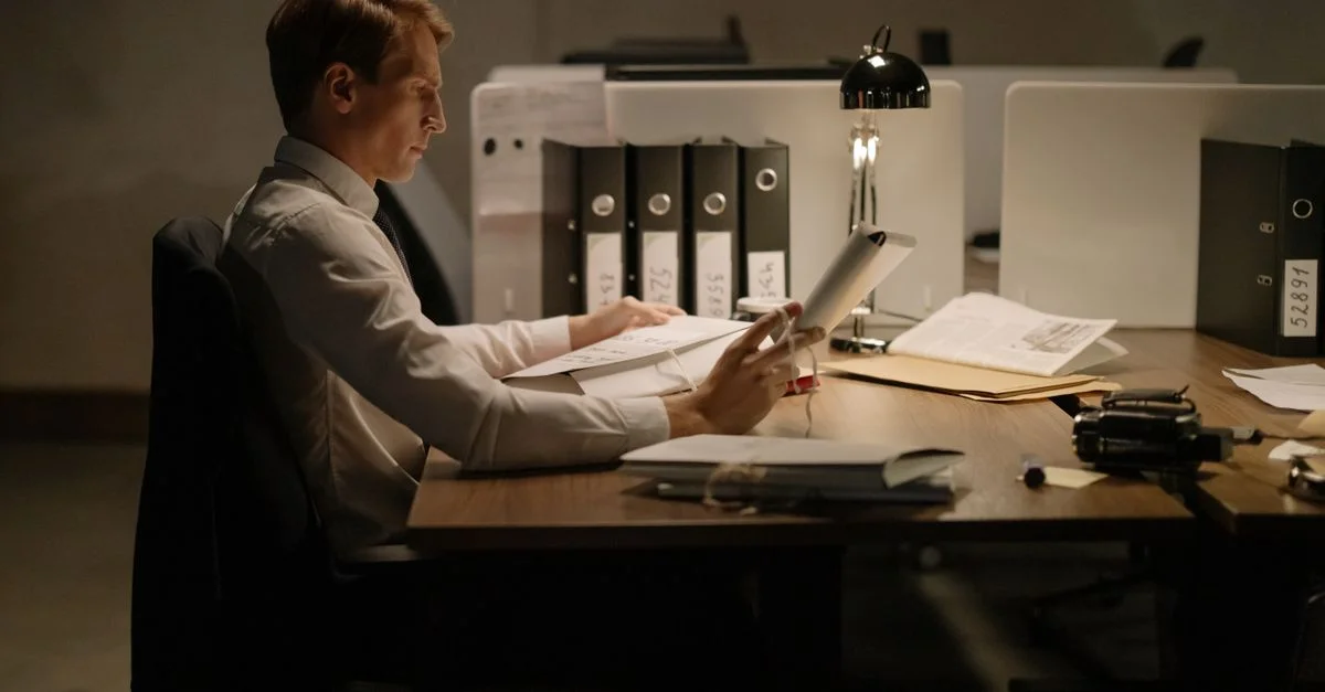 Un employé de bureau en tenue formelle, intensément concentré sur la lecture de documents à un bureau avec des dossiers et une lampe.