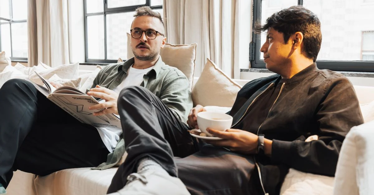 Deux hommes assis sur un canapé confortable, l'un lisant un magazine, en train de déguster un café dans un salon lumineux.