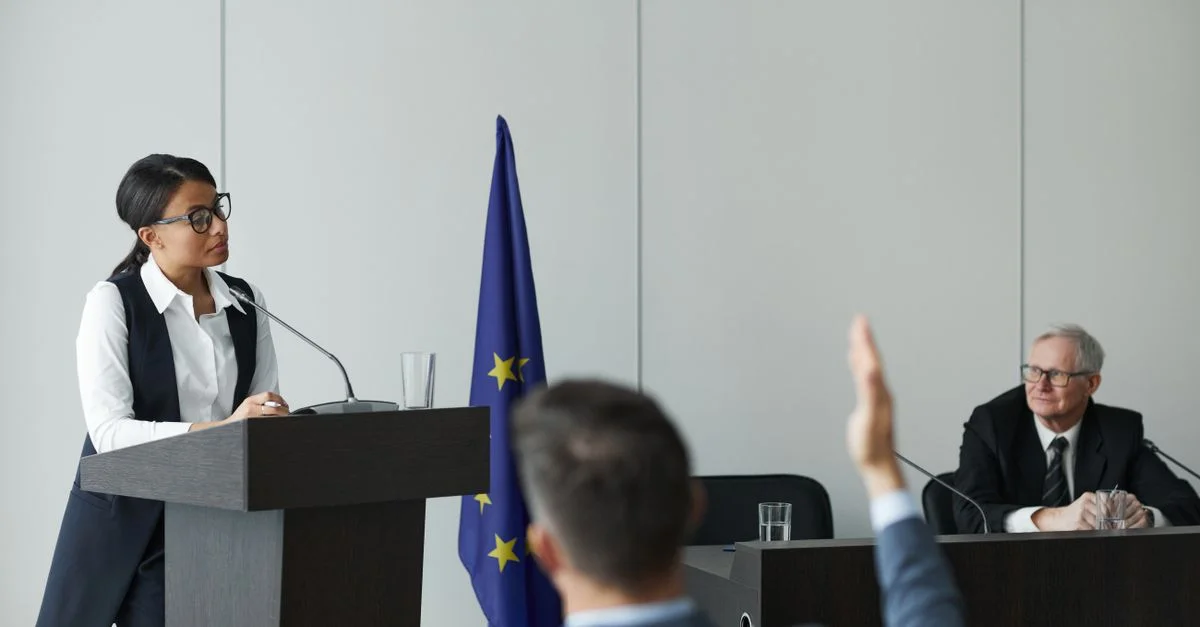 Une femme s'exprimant lors d'une conférence de l'Union européenne pendant que des délégués écoutent attentivement.