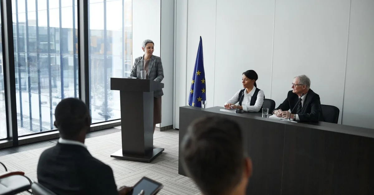 Environnement de conférence professionnel avec une oratrice au podium et un public attentif dans une salle de réunion moderne.