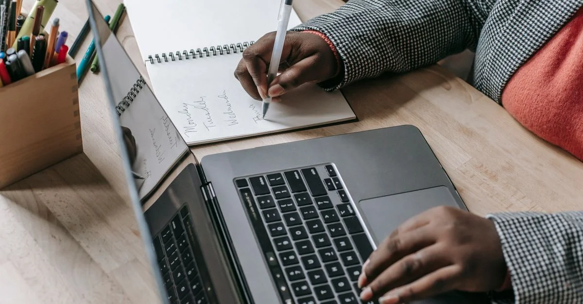 Femme concentrée prenant des notes tout en utilisant un ordinateur portable, mettant en avant le multitâche professionnel et un environnement de travail moderne.